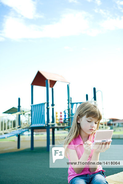 Mädchen sitzt auf dem Spielplatz und spielt mit einem tragbaren Videospiel.