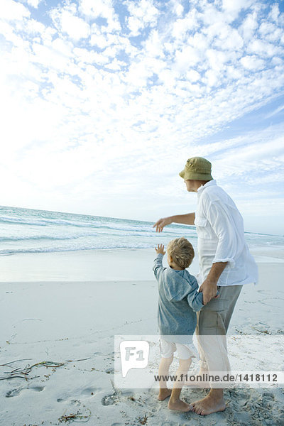 Älterer Mann steht mit Enkel am Strand und zeigt in die Ferne.
