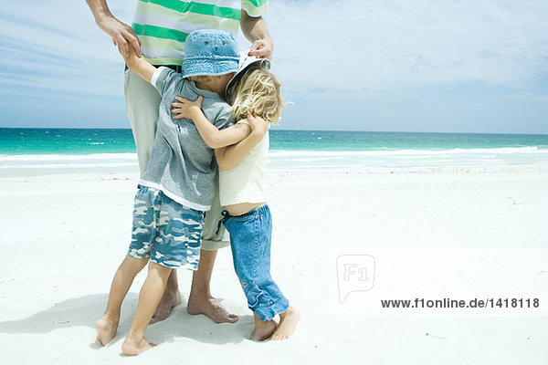 Mann am Strand stehend mit Sohn und Tochter  die seine Beine umarmen  Taille unten