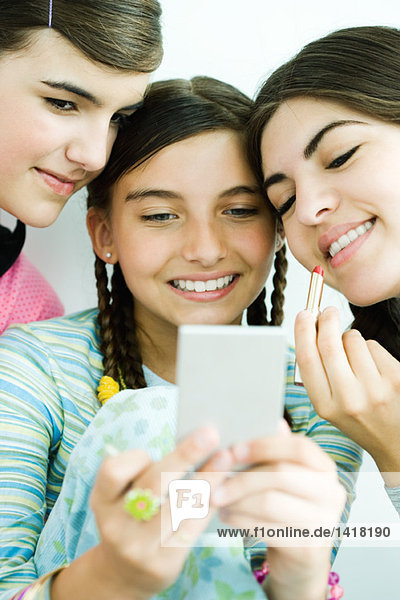 Drei junge Freundinnen schauen sich im Handspiegel an  eine hält Lipgloss.
