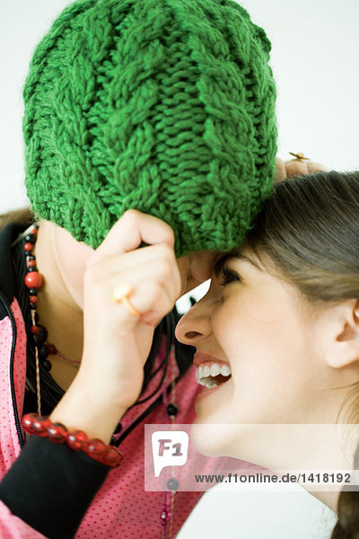 Two young female friends with heads together  one pulling knit hat over eyes while the other laughs