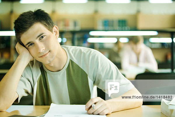Männlicher Student sitzt in der Bibliothek und lächelt vor der Kamera.