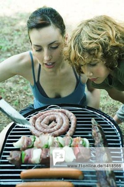 Junge Frau und Junge beugen sich über das Barbecue und schauen sich Fleisch an.
