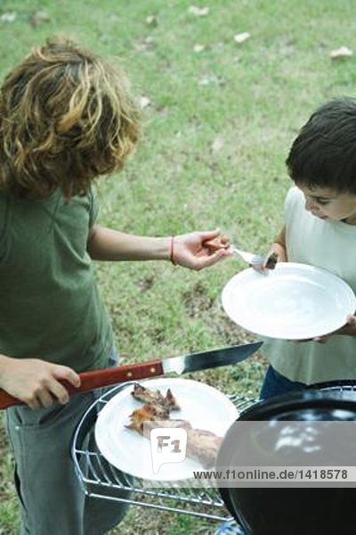 Zwei Jungs stehen neben dem Grill  einer legt ein Stück Fleisch auf den Teller des anderen.