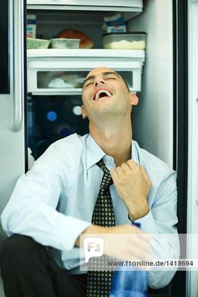 Mann sitzt vor offenem Kühlschrank  zieht am Kragen  Kopf zurück