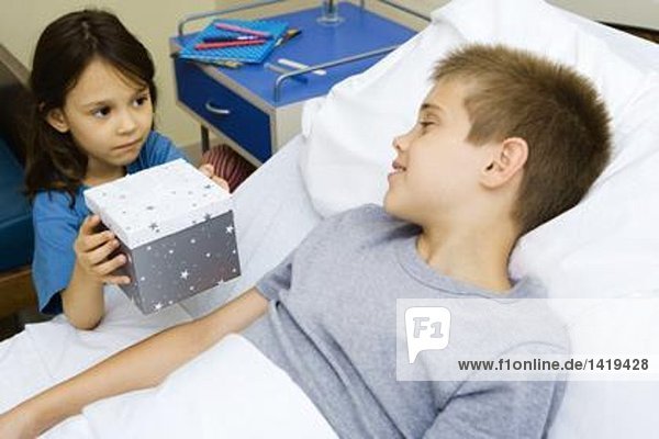 Junge liegt im Krankenhausbett und bekommt ein Geschenk von einem Mädchen.