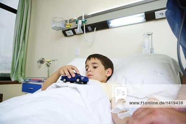 Junge liegt im Krankenhausbett und hält Spielzeug.