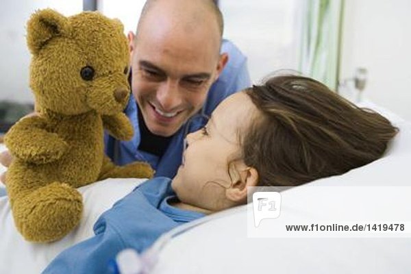 Mädchen im Krankenhausbett liegend  Krankenschwester hält Teddybär hoch