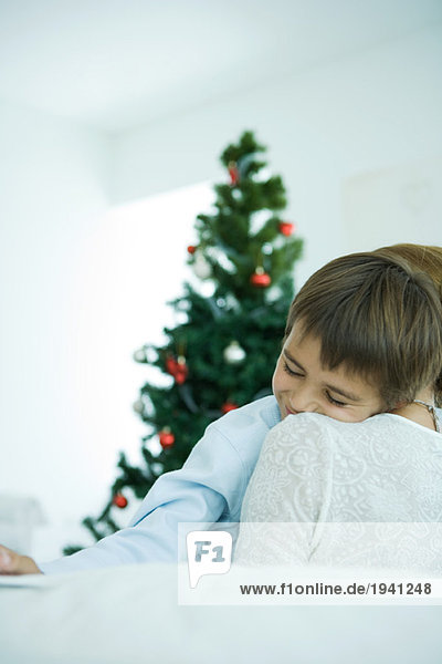 Mutter sitzt auf dem Sofa  hält den Jungen in den Armen  Weihnachtsbaum im Hintergrund  Junge lächelnd