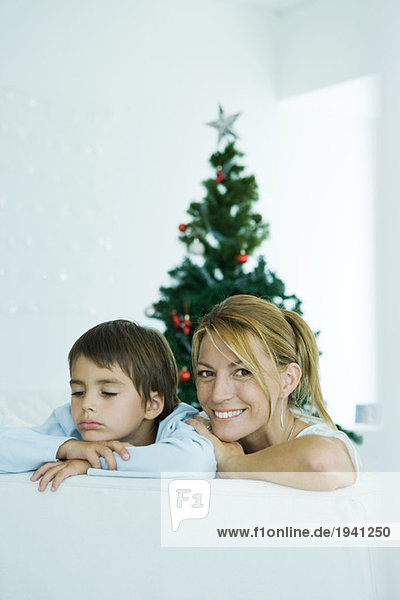 Junge und Mutter auf dem Sofa  Weihnachtsbaum im Hintergrund  Frau lächelt in die Kamera  Junge runzelt die Stirn