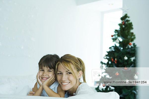 Mädchen und Mutter auf dem Sofa lächelnd vor der Kamera  Weihnachtsbaum im Hintergrund