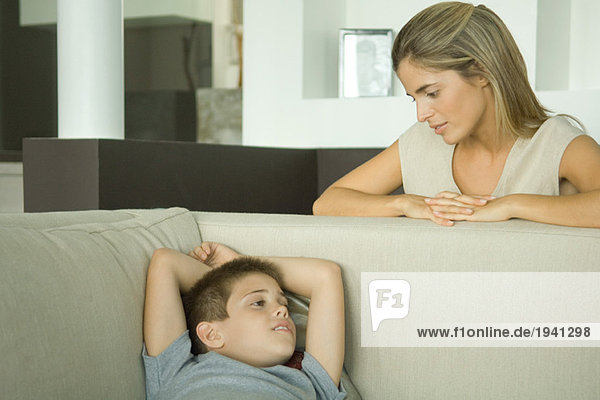 Boy lying on sofa talking to mother looking down at him
