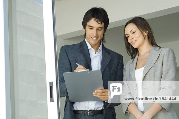 Männlicher Immobilienmakler  der dem weiblichen Kunden das Haus zeigt und auf die Zwischenablage schreibt.