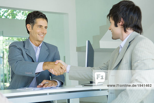 Junger Mann schüttelt dem reifen Geschäftsmann die Hand über den Schreibtisch  lächelnd