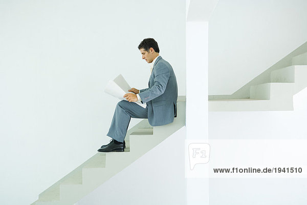 Mann im Anzug sitzt auf einer Treppe und sieht sich Dokumente an.