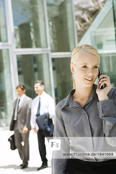 Young businesswoman using cell phone outdoors  looking away  businessmen walking in background