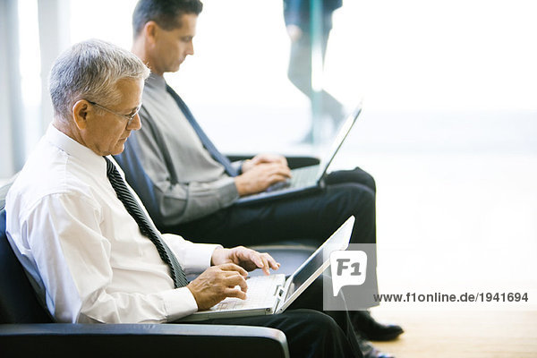 Two businessmen sitting in lobby  both using laptop computers  side view