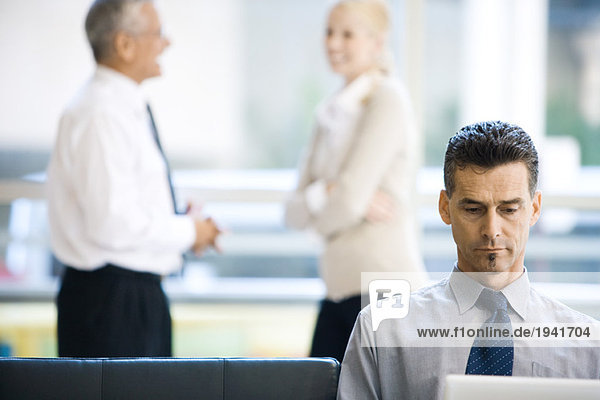Businessman sitting  using laptop computer  associates standing in background