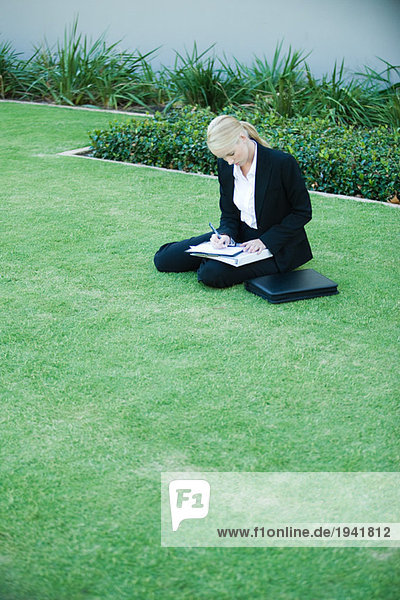 Young businesswoman sitting on the ground outdoors  writing on clipboard