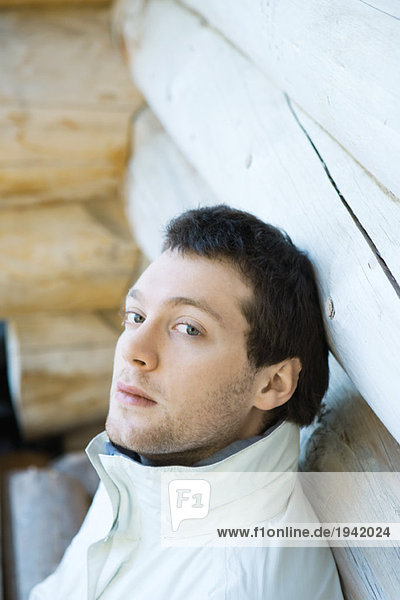 Young man leaning against wall of log cabin  looking at camera