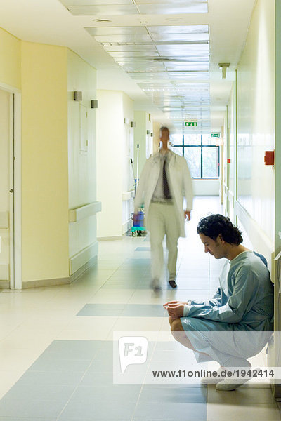 Male patient crouching in hospital corridor  looking down  doctor walking in background