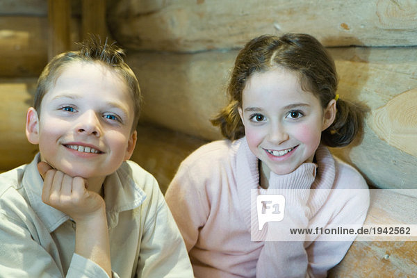 Junge und Mädchen sitzen Seite an Seite  Hand unter dem Kinn  lächeln vor der Kamera