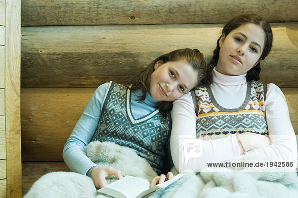 Zwei Teenagermädchen teilen sich eine Decke  lächeln in die Kamera  eine hält das Buch  die andere ruht mit dem Kopf auf der Schulter.