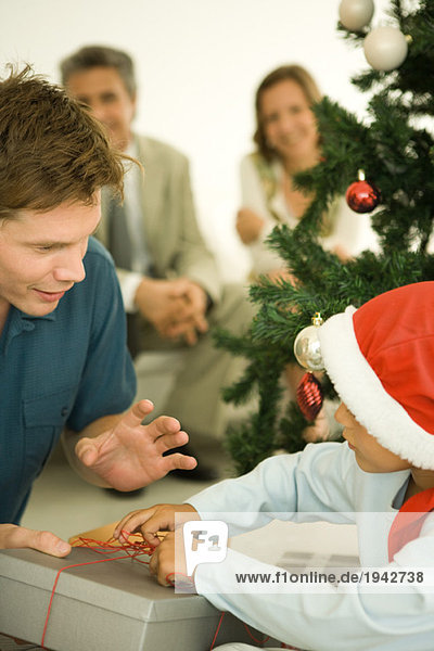 Vater und Sohn eröffnen Weihnachtsgeschenk vor dem Weihnachtsbaum  Junge mit Weihnachtsmütze