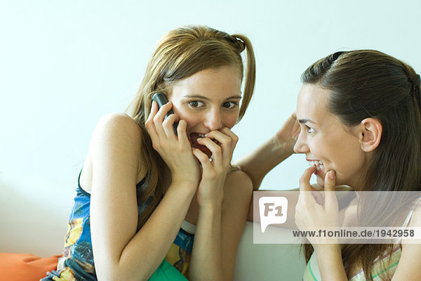 Two young friends sitting together  smiling  one using cell phone  covering mouth