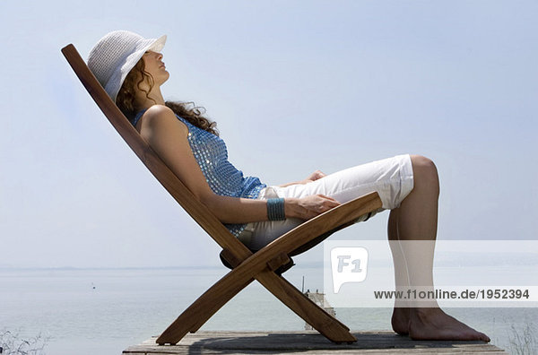 Junge Frau sitzt auf Liegestuhl am Steg  Seitenansicht