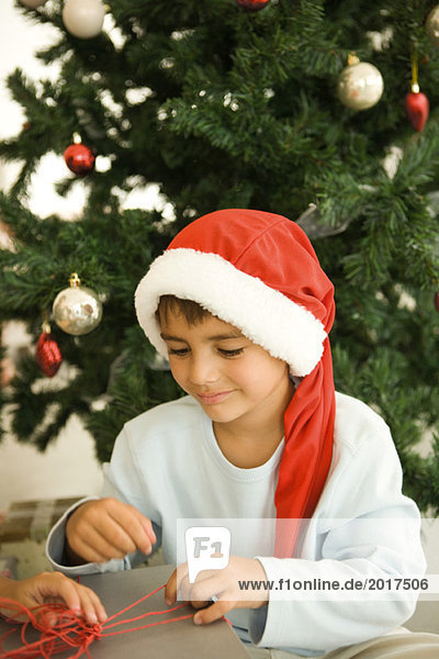 Junge Eröffnungsgeschenk vor dem Weihnachtsbaum  mit Weihnachtsmütze  lächelnd
