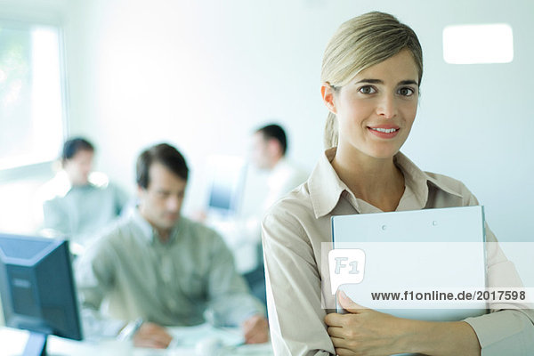 Young businesswoman holding laptop in front of chest in office  smiling at camera  portrait