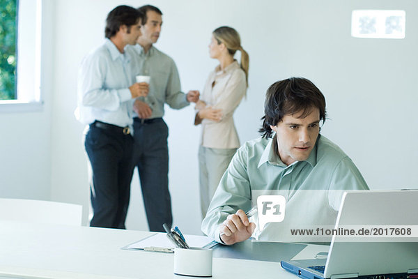 Businessman working on laptop  colleagues standing in background