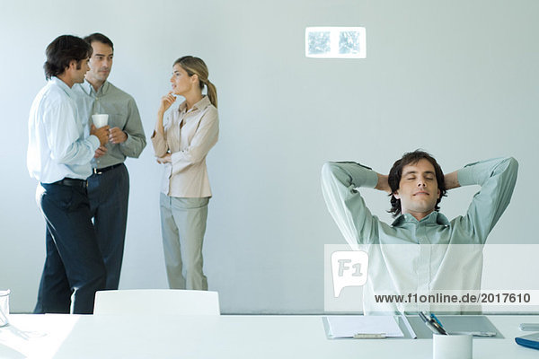 Geschäftsmann am Schreibtisch mit geschlossenen Händen hinter Kopf und Augen  Kollegen im Hintergrund