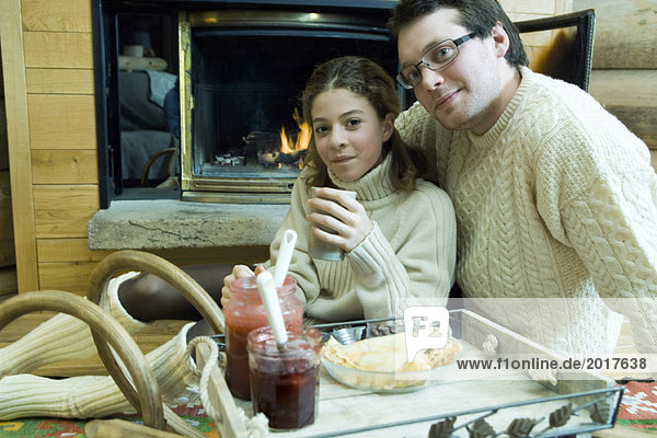 Young man and teen girl drinking hot drinks by fire place