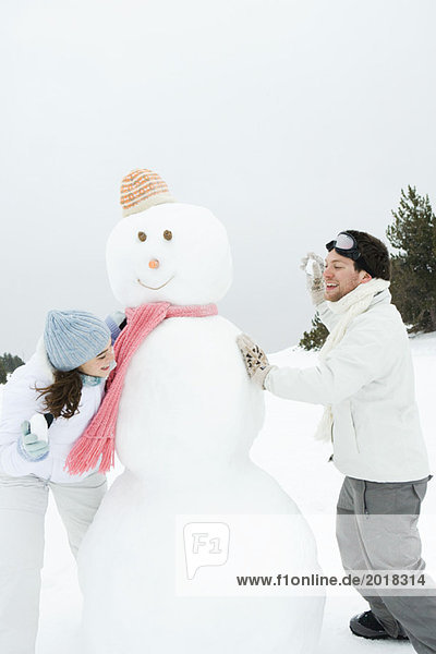 Young couple peeking around snowman at each other  holding up snowballs