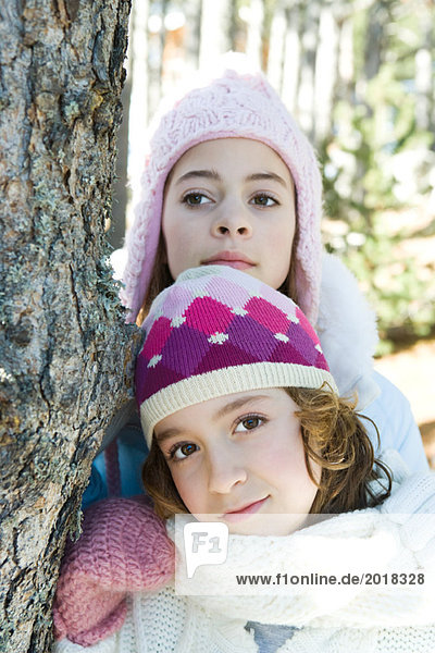 Two young friends leaning against tree trunk  dressed in winter clothing  one looking at camera