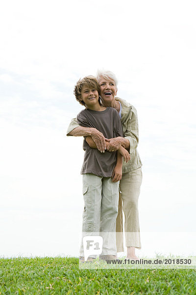 Seniorin und Enkelin auf Gras stehend  Frau umarmt den Jungen von hinten  lächelnd und aus dem Rahmen schauend  volle Länge
