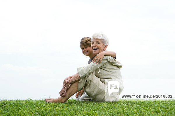 Seniorin und Enkelin auf Gras sitzend  lächelnd vor der Kamera  volle Länge