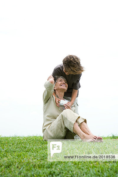 Seniorin auf Gras sitzend  Enkelin lehnt sich über die Schulter  überreicht ihr Geschenk  volle Länge