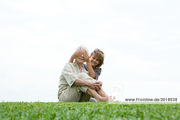 Seniorin auf Gras sitzend  Enkelin  die ihre Augen von hinten bedeckt  lächelnd vor der Kamera  volle Länge