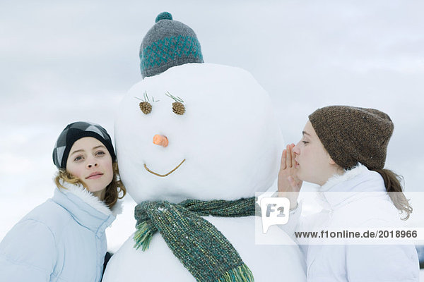 Teen girls on either side of snowman  one whispering  the other listening