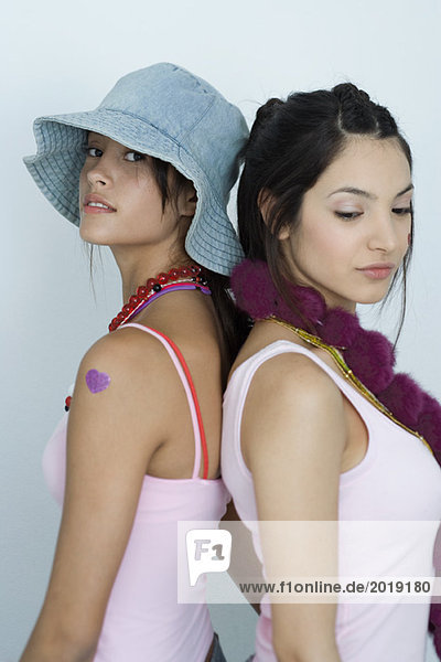 Zwei junge Freundinnen  Rücken an Rücken stehend  eine mit Blick auf die Kamera  die andere mit Blick nach unten  Taille hoch  Porträt