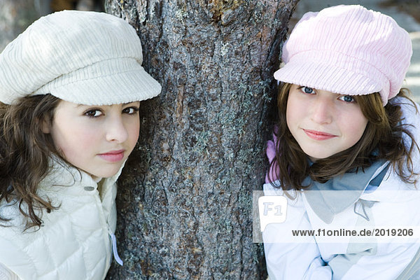 Two young friends leaning against tree trunk  both wearing caps  smiling at camera  portrait