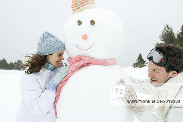 Two young friends peeking around snowman at each other  smiling