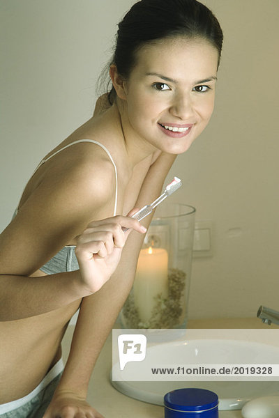 Junge Frau steht am Waschbecken  hält Zahnbürste  lächelt in die Kamera