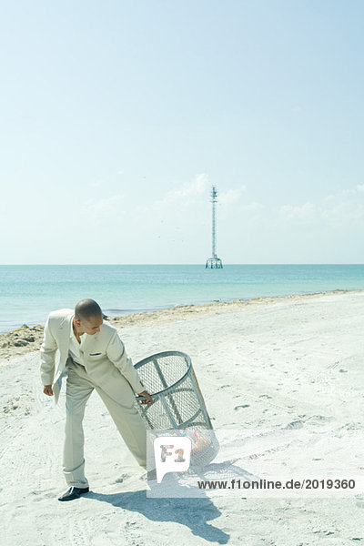 Man in suit pulling garbage can across sunny beach  looking back over shoulder  full length