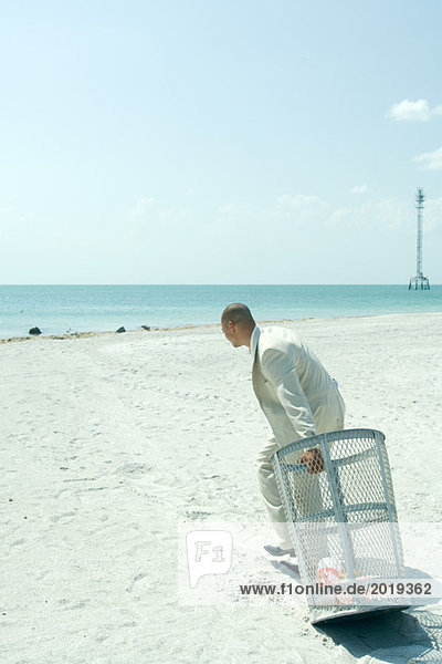 On beach  man in suit pulling garbage can towards ocean  side view  full length