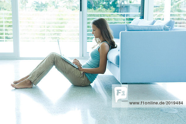 Teenage girl leaning against sofa  using laptop computer  full length
