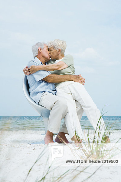 Senior couple sitting in chair together on beach  hugging and kissing  woman sitting on man's lap  full length